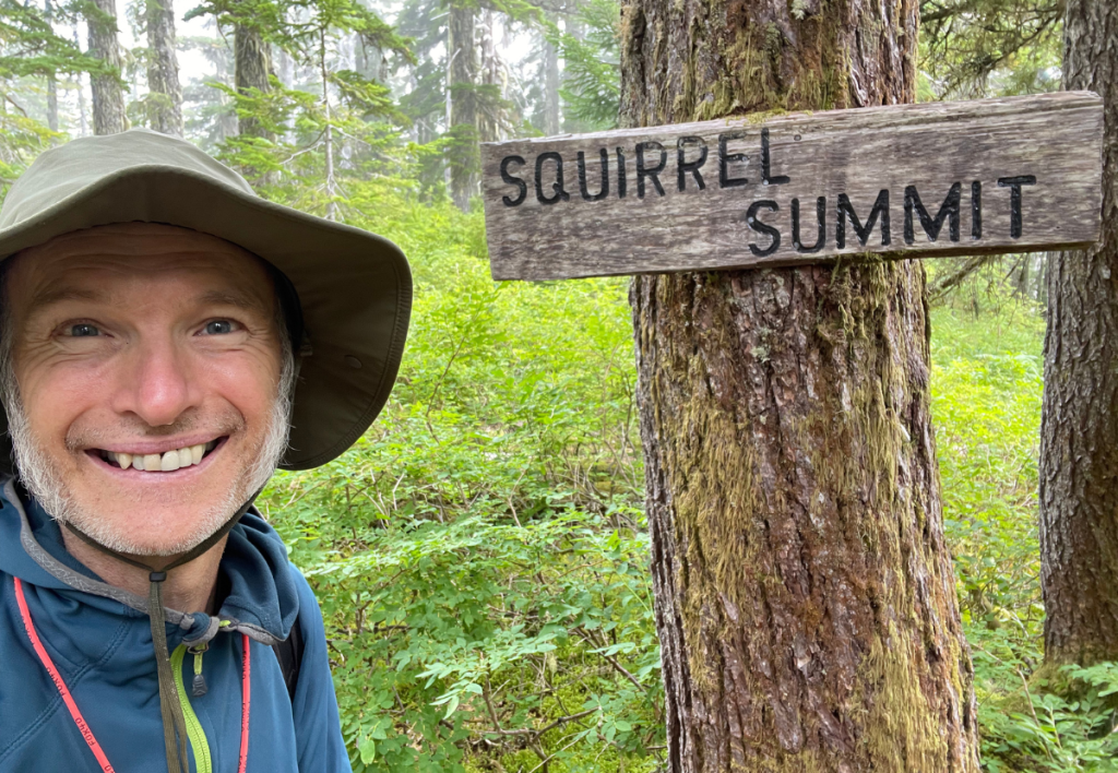arne-pedersen-hypnotherapy-victoria-kludahk-summit-sign-squirrel-mindfulness-joy Arne Pedersen smiling beside a “Summit” sign and squirrel figure on the Kludahk Trail—symbolizing joy, mindful milestones, and transformational healing.