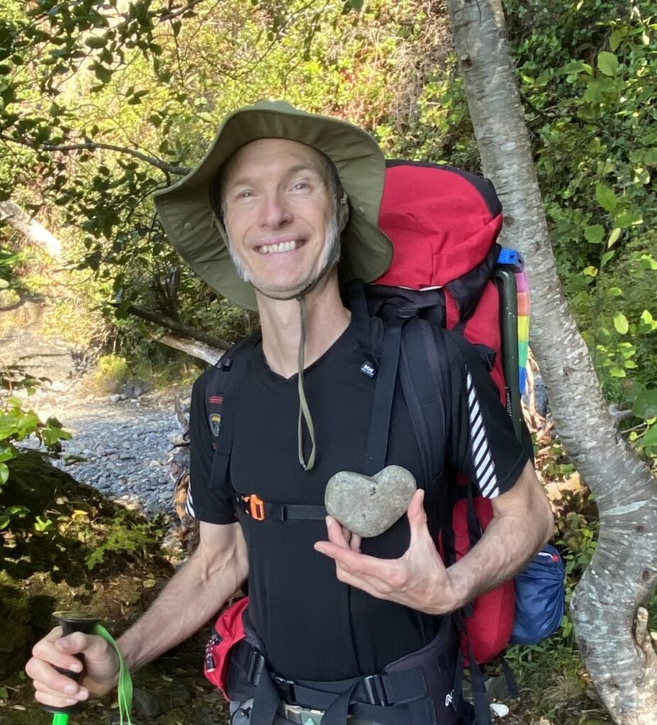 Arne Pedersen smiling outdoors while holding a heart-shaped rock, representing warmth, support, connection, and positive energy.